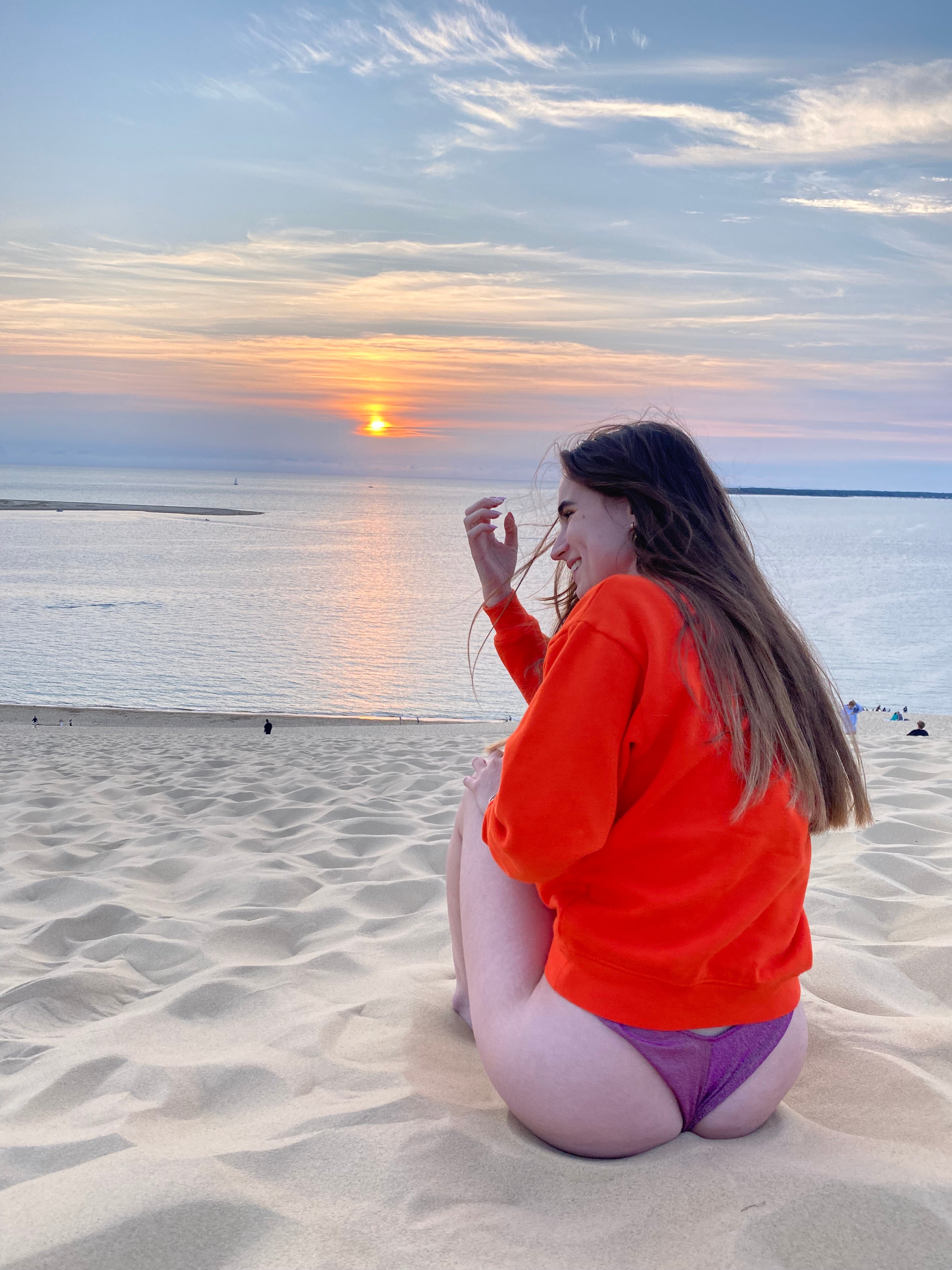 Femme portant un sweat-shirt orange et un bas de maillot de bain violet, assise sur une dune de sable avec une vue magnifique sur le coucher de soleil sur l'océan. Moment de détente à la plage.